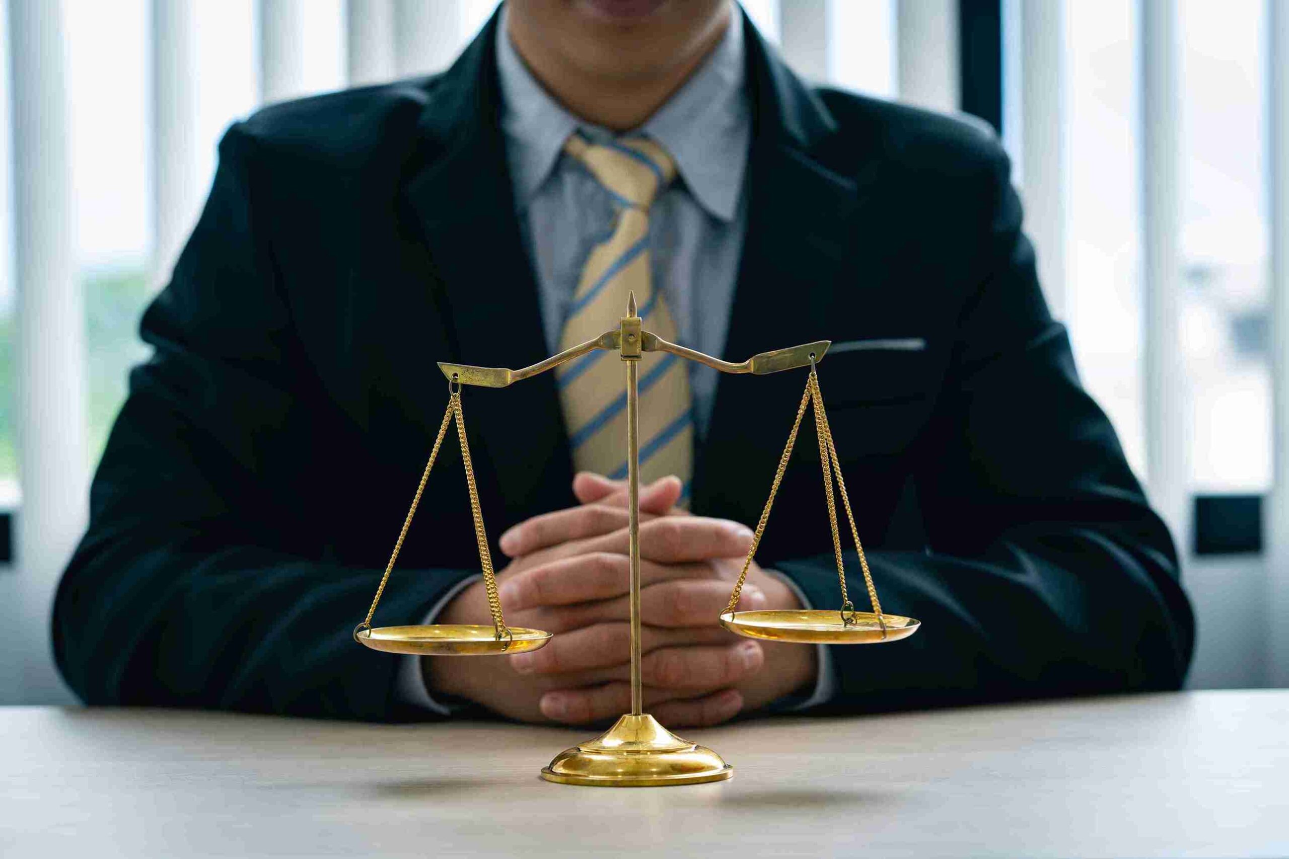 A person in a suit sits at a desk with hands clasped behind a golden balance scale, symbolizing justice, in an office setting.