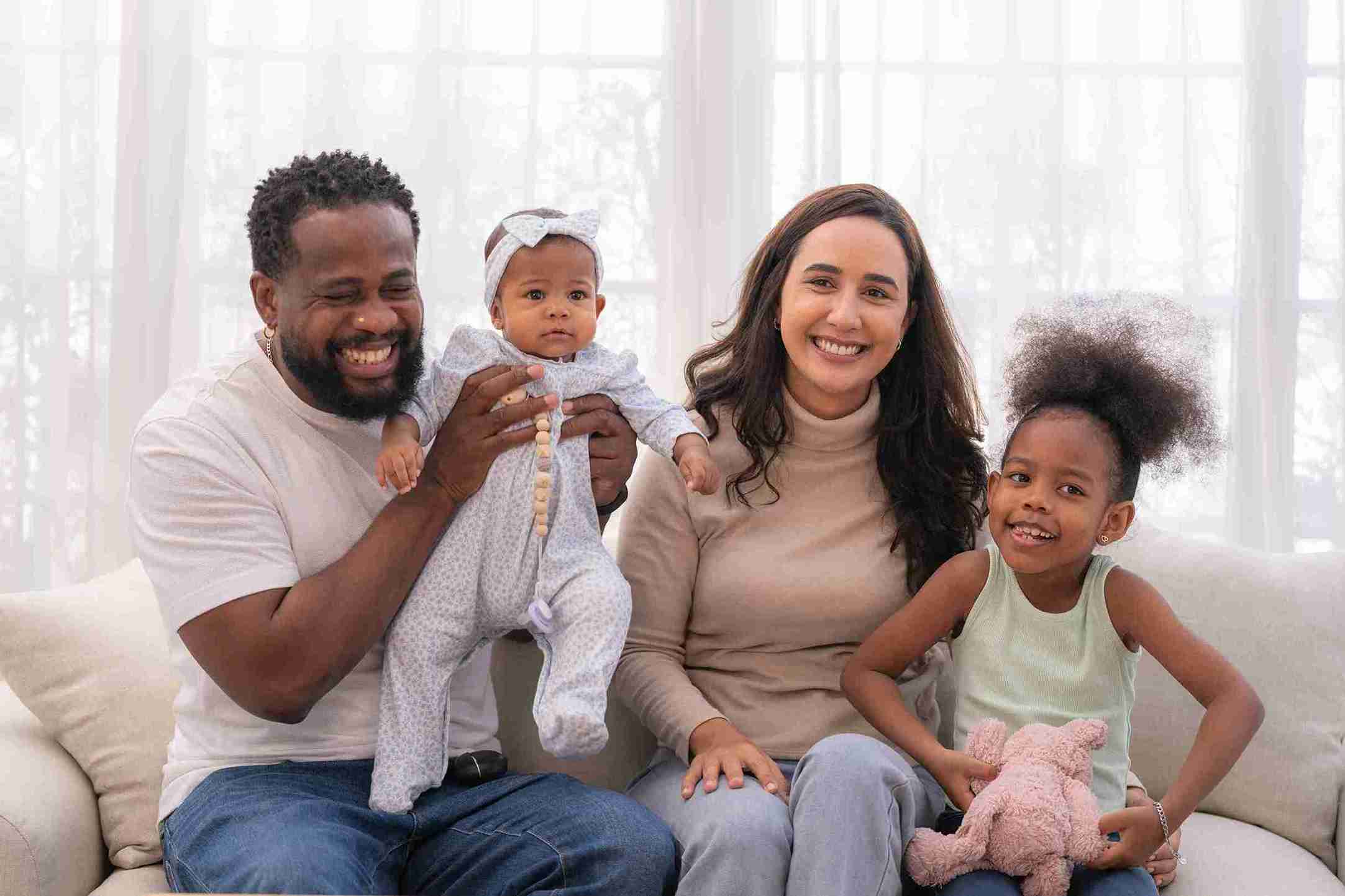 A family of four, including two children, happily sitting together on a cozy couch in a warm living room.