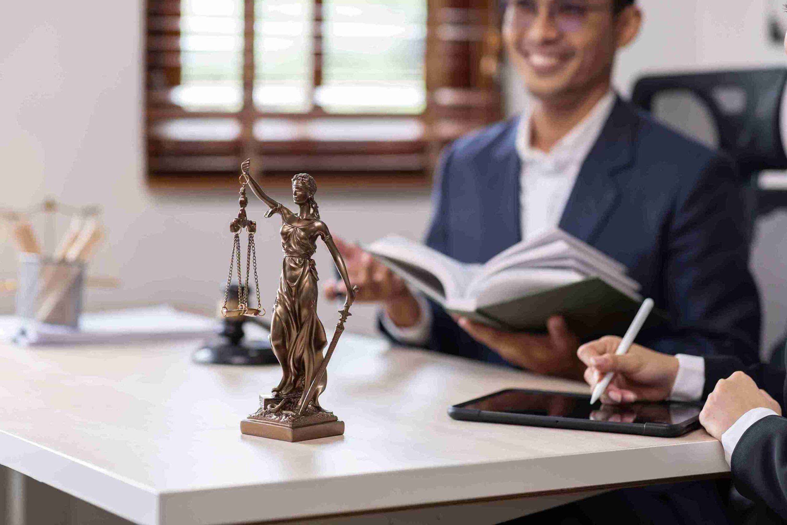 A man and woman sit at a table with a tablet and a statue of Lady Justice between them, discussing legal matters.