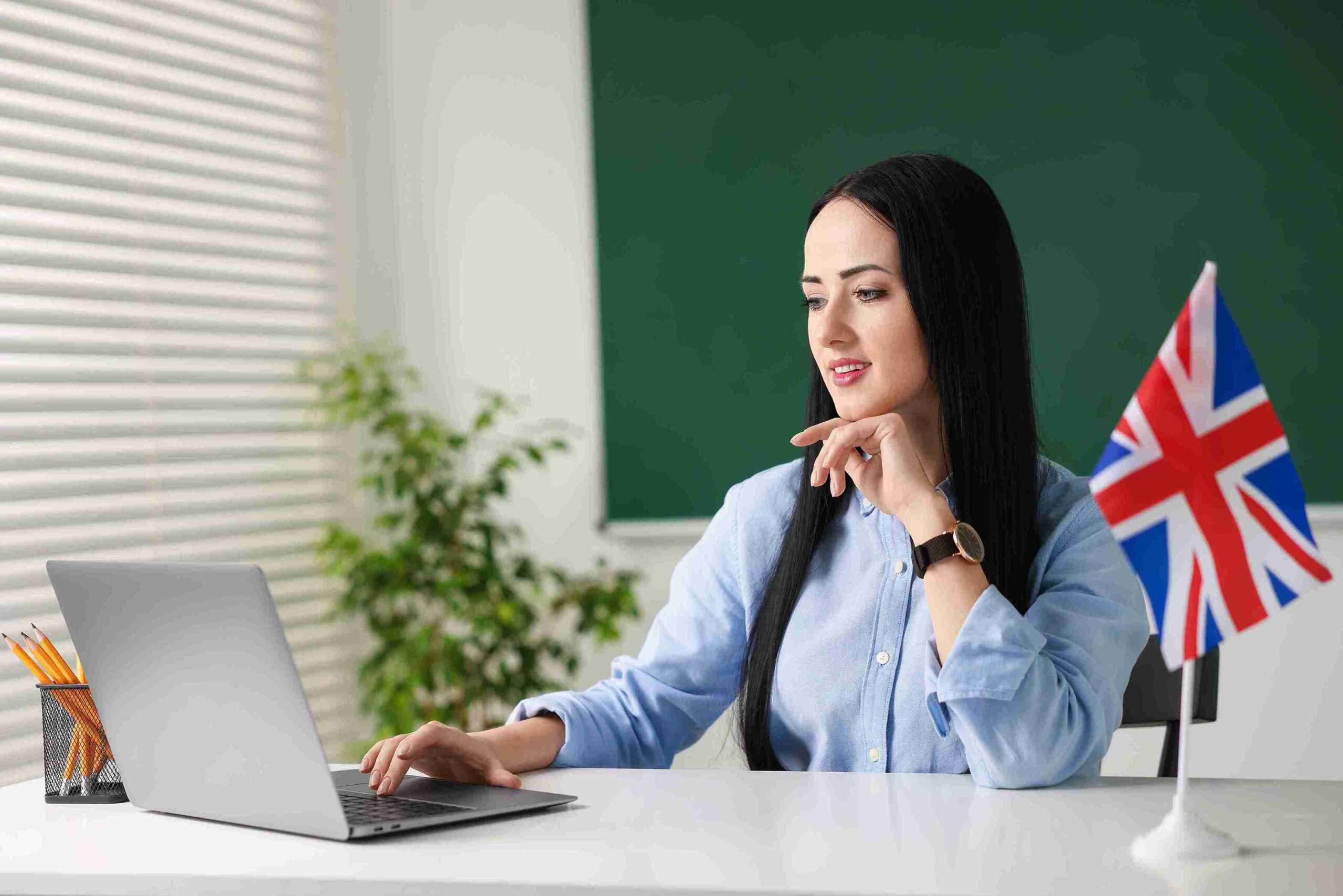A woman in a classroom with a laptop, sitting next to a British flag, engaged in her studies.
