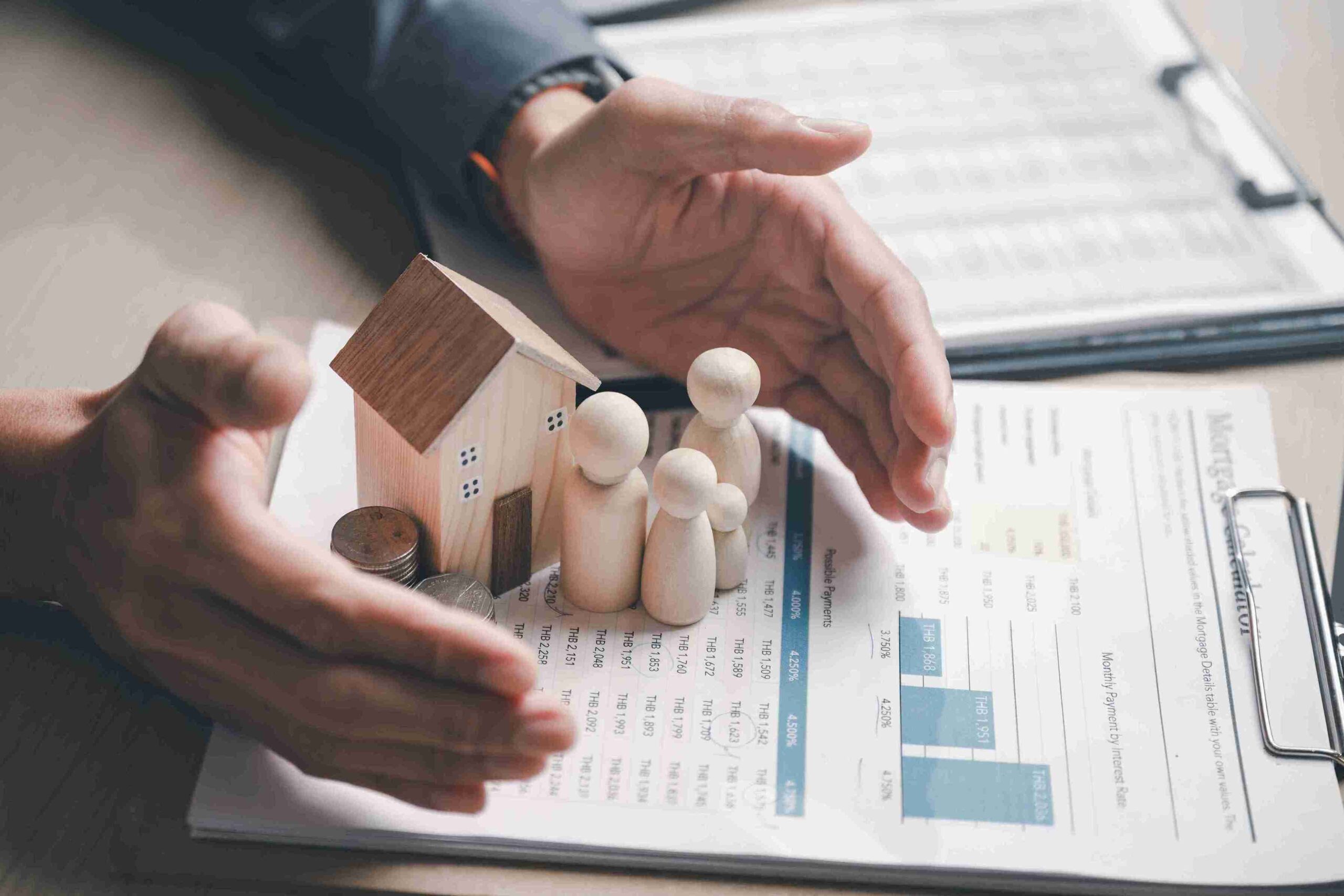 Hands encircle wooden figures of a family and a house on charts and documents. The image conveys financial security and protection.
