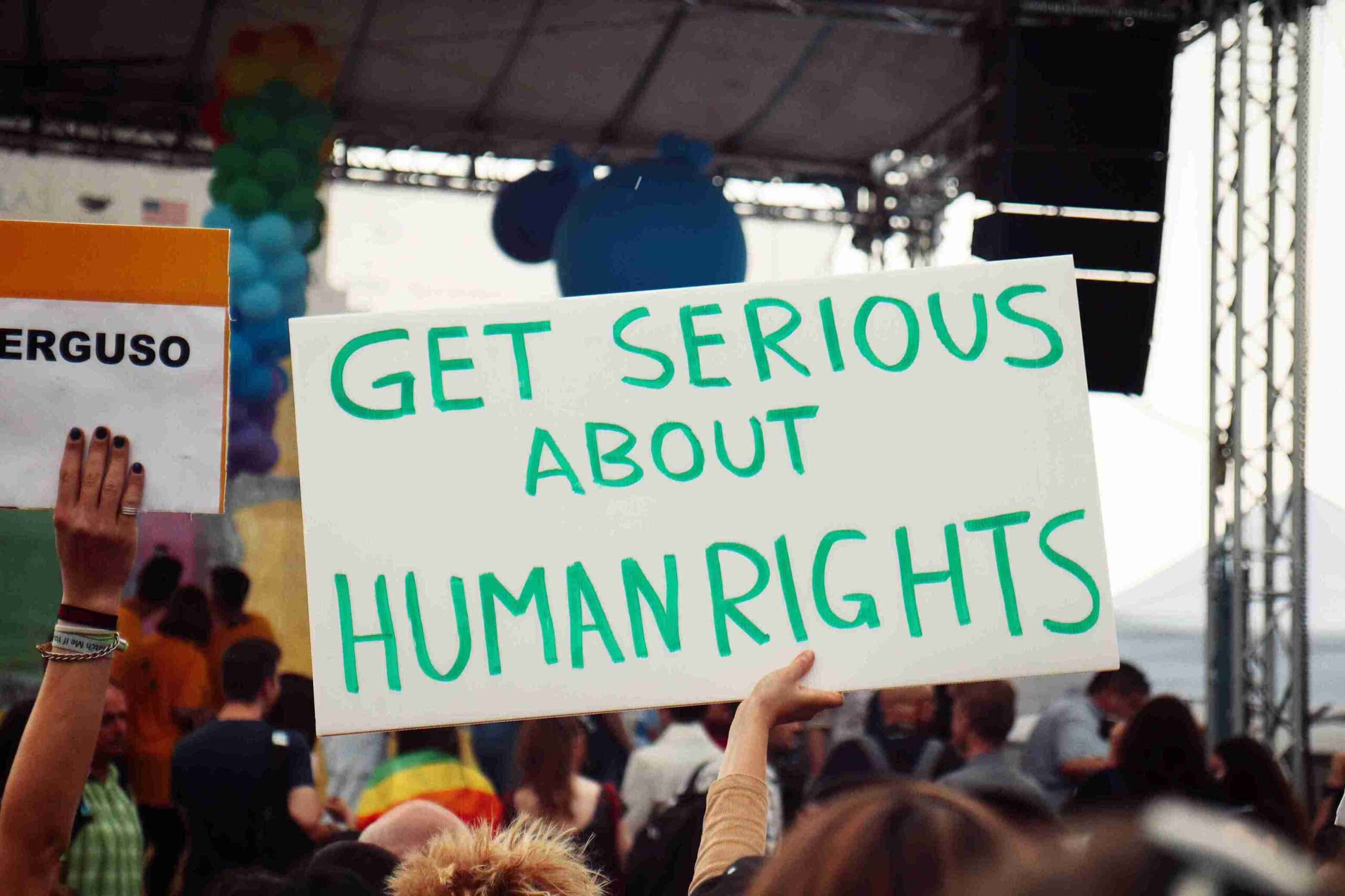 A diverse crowd holds a sign reading "Get Serious About Human Rights," advocating for social justice and equality.