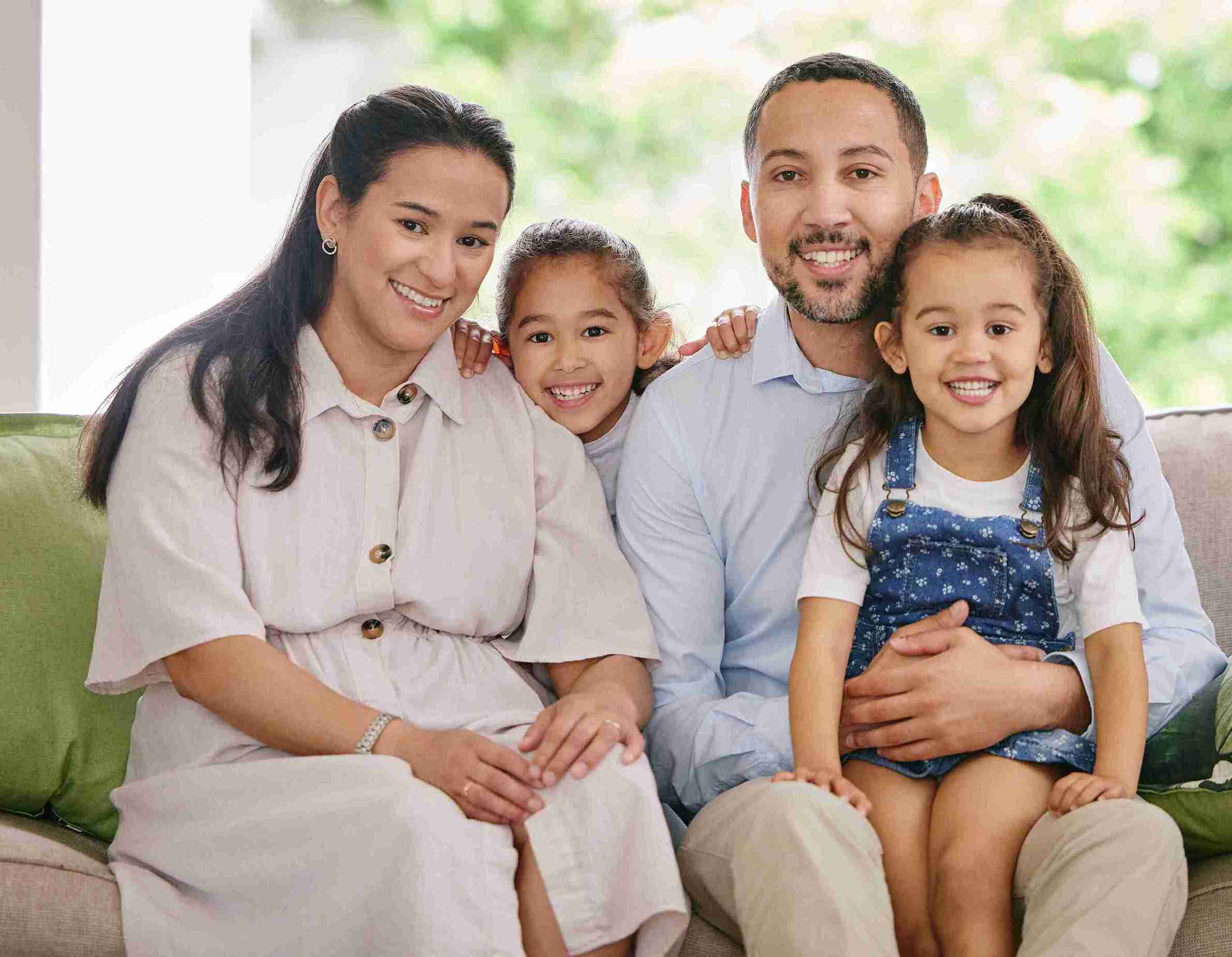 A family of four sits together on a couch, smiling at the camera, with a baby nestled in the mother's arms.