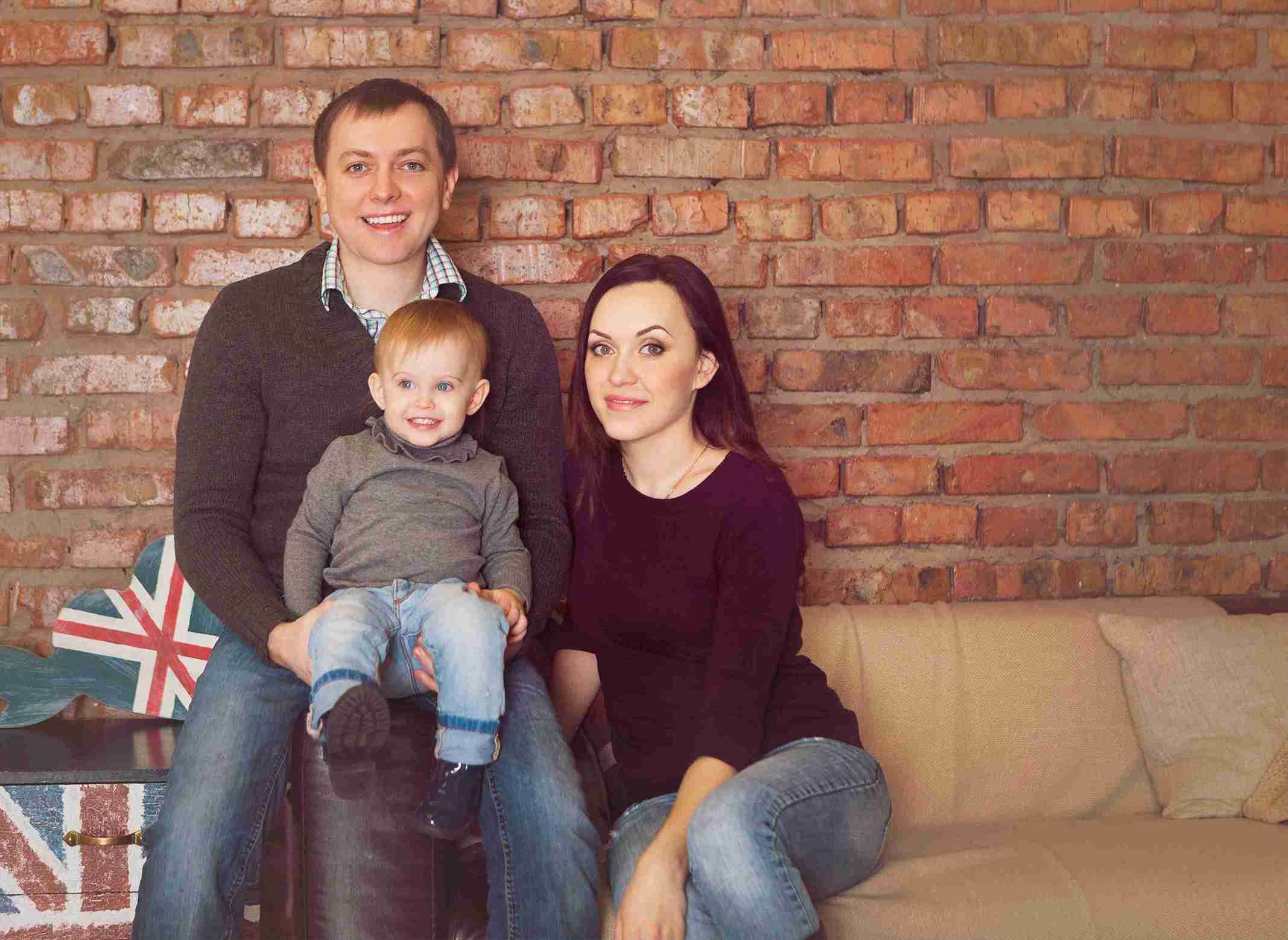 A man and woman sit on a couch with a child, all smiling, in a cozy living room setting.