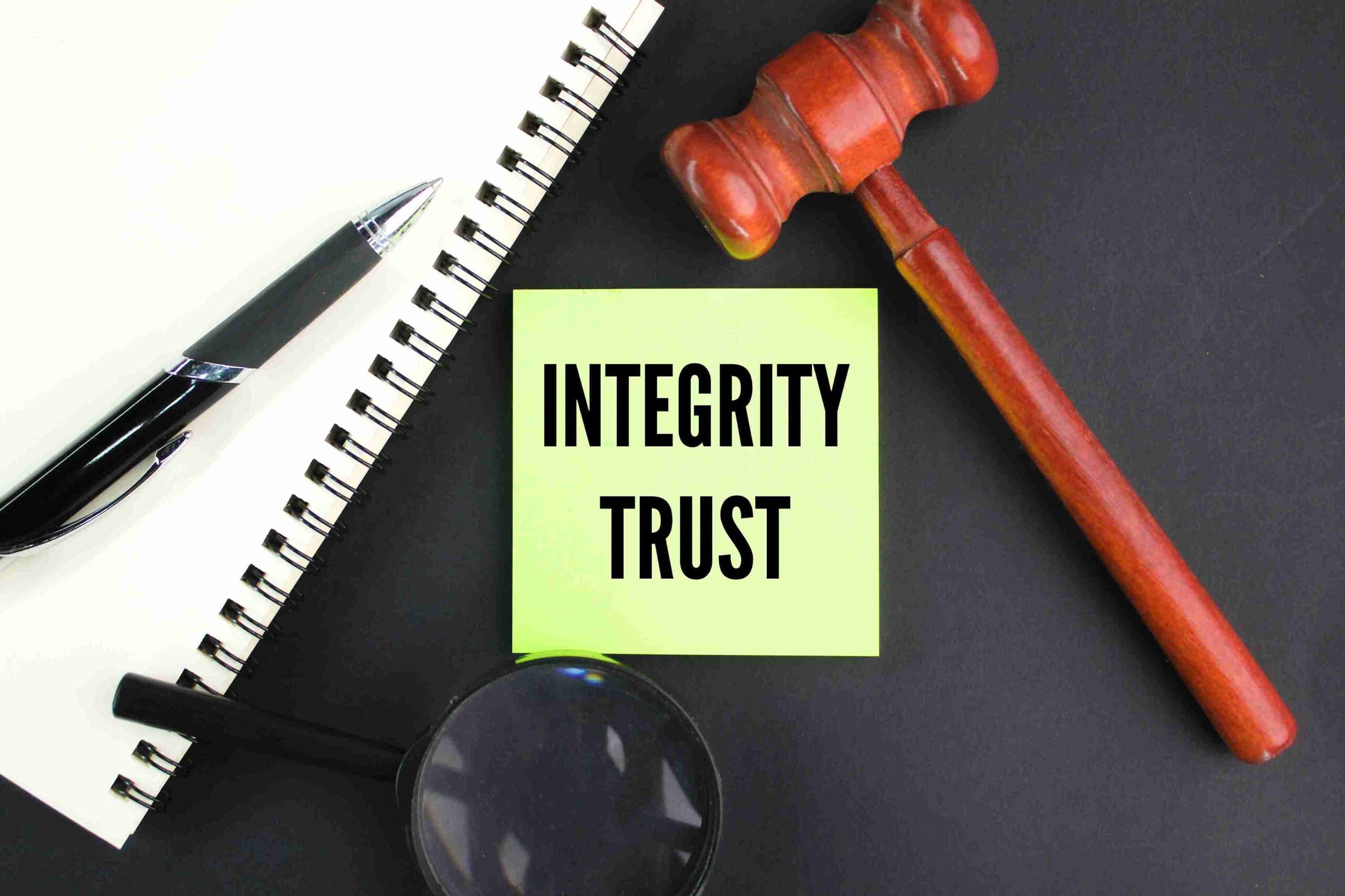 A wooden gavel, a notepad with a pen, a magnifying glass, and a sticky note with integrity trust on a dark surface, conveying Trusts & Waqf Services.
