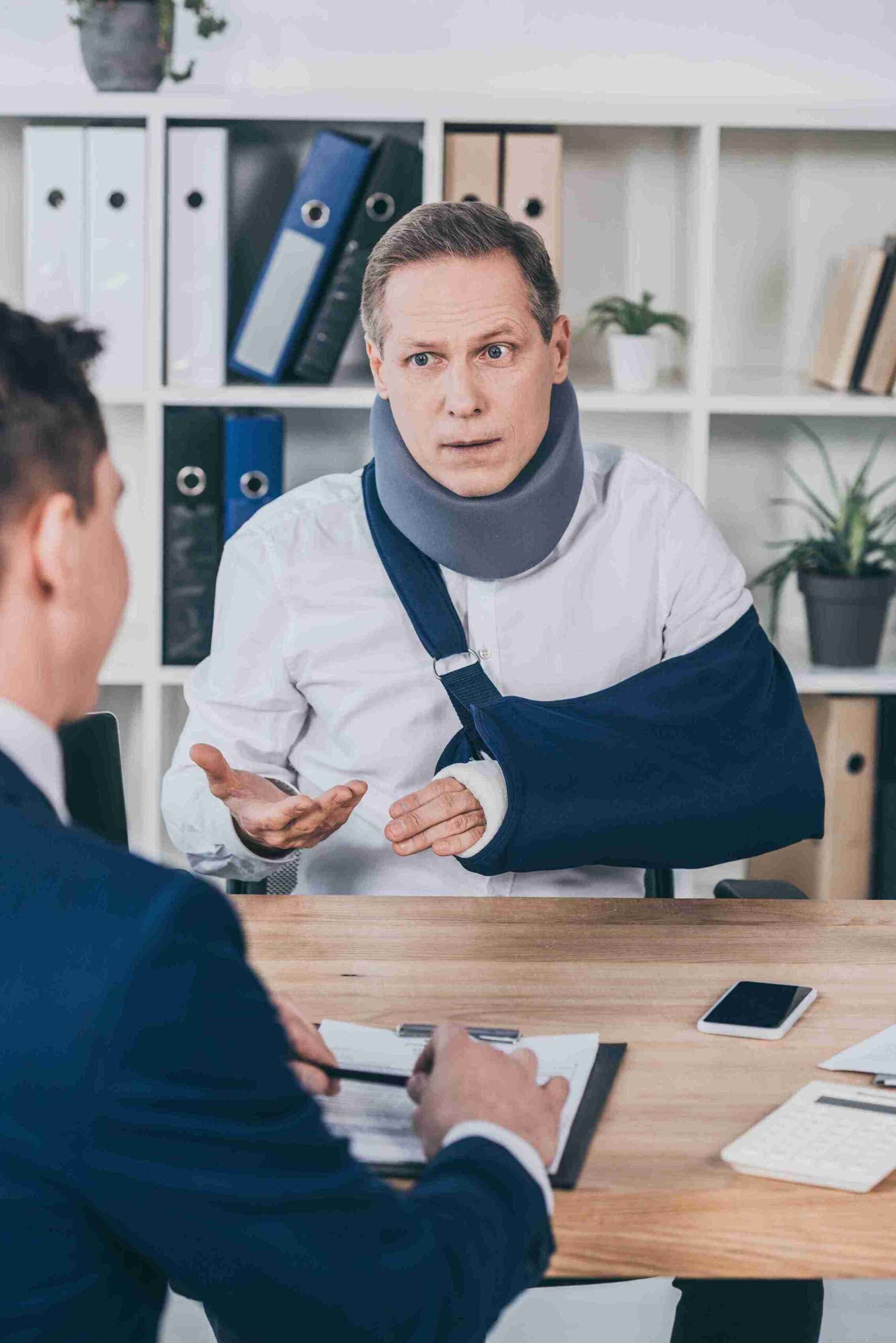 A man in a neck brace sits at a table with another man, discussing potential recovery of financial losses.