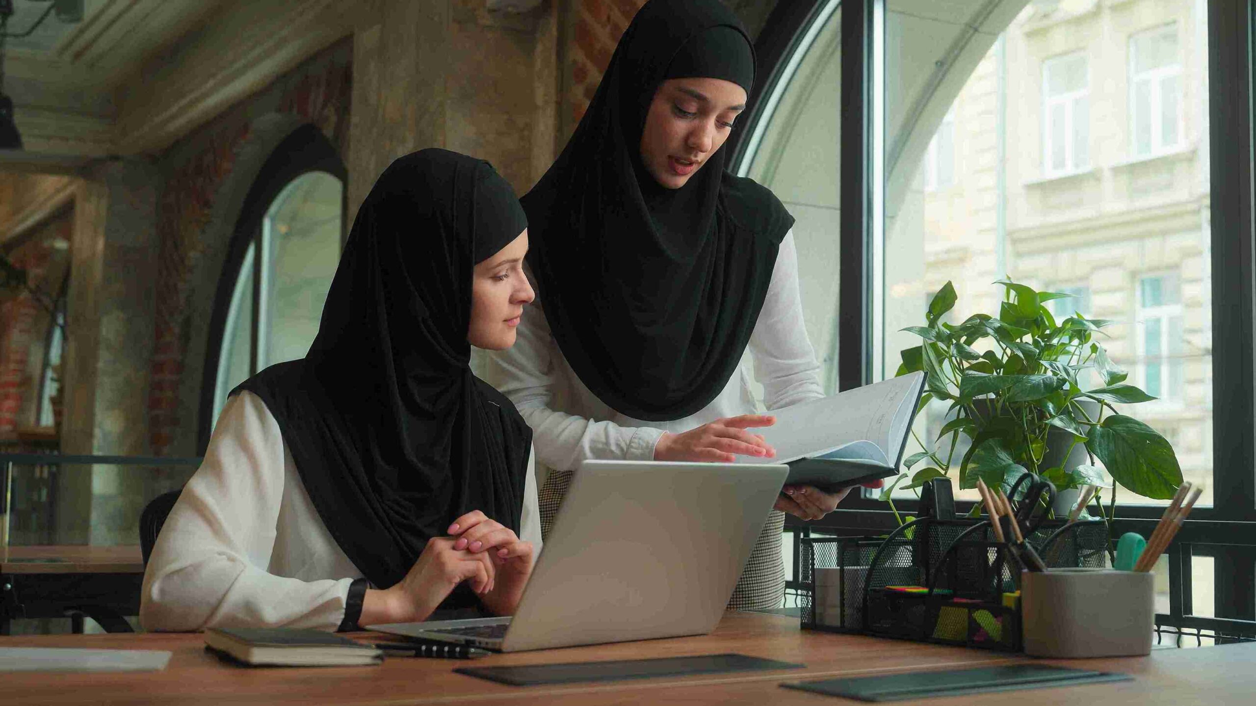 Two women in hijabs working in modern office with laptop and notebook, representing balancing UK law with Shariah principles for legally arrangements.