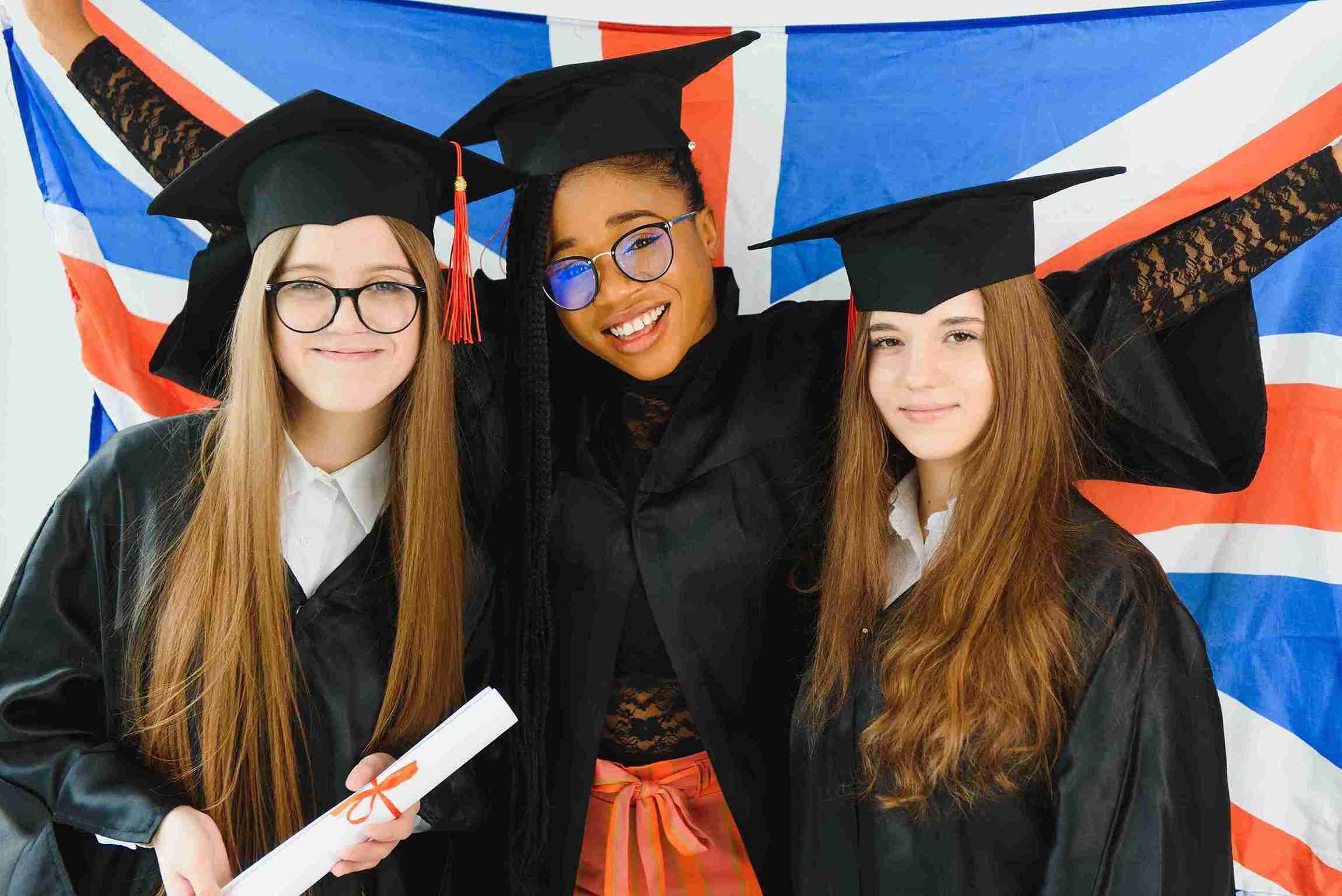 Three young women in graduation gowns and caps proudly pose with a Union Jack flag, celebrating their achievement.