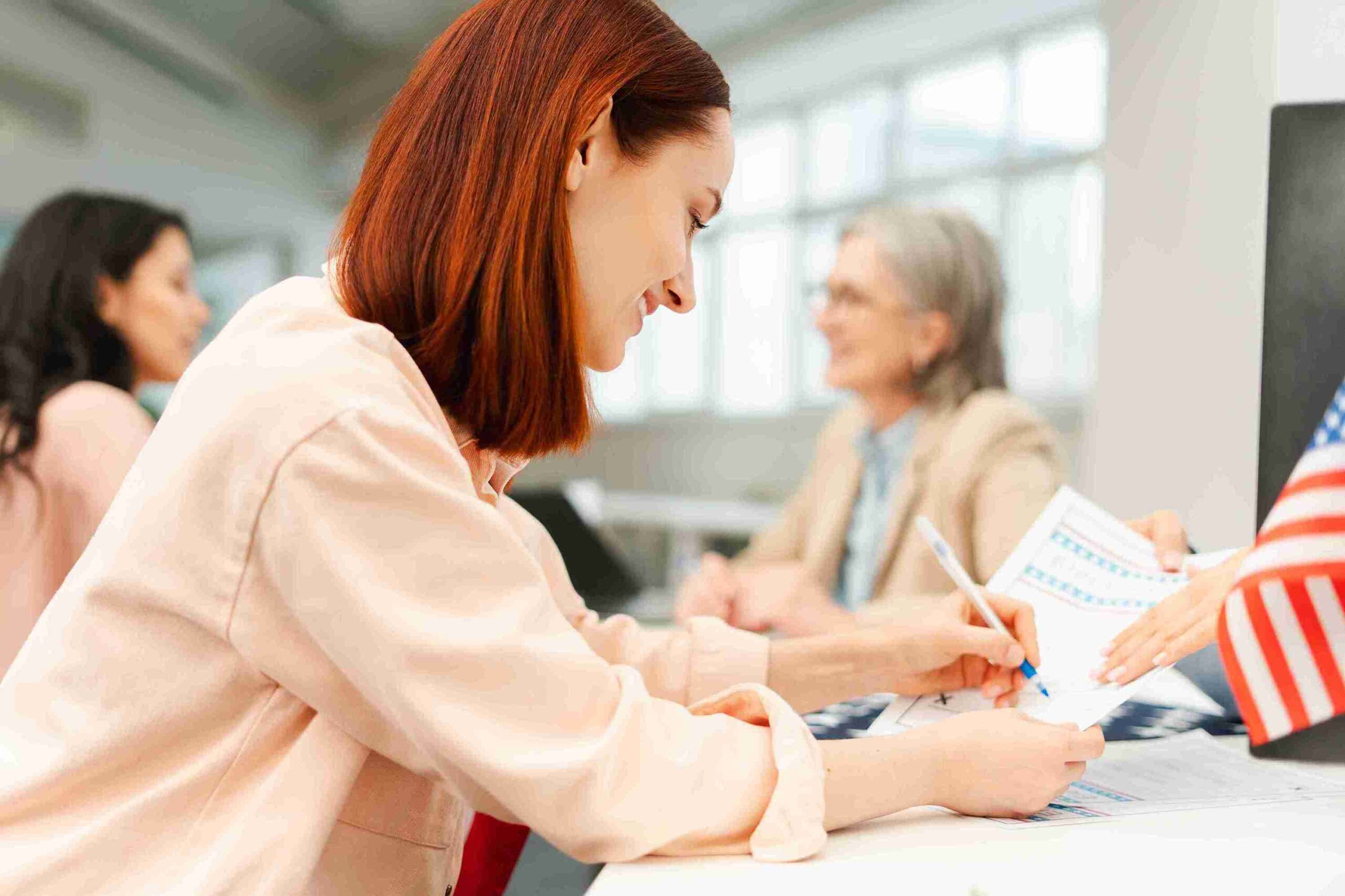A woman signs documents at a desk, with an American flag displayed prominently in the background.