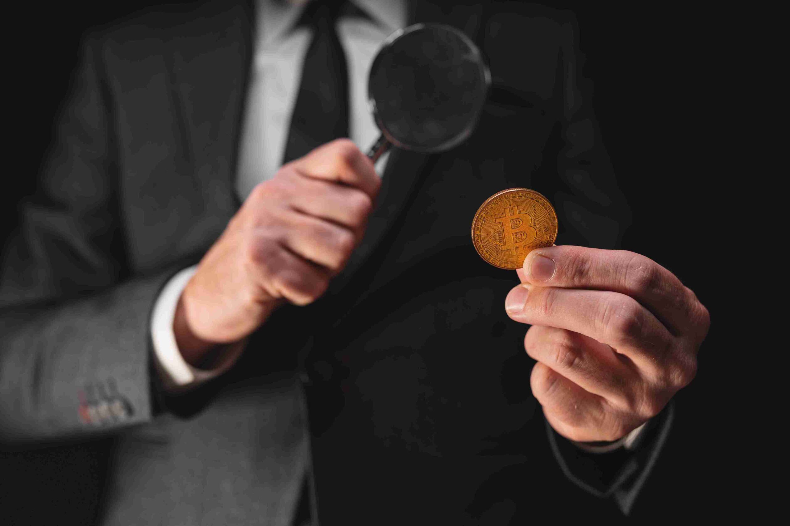 A man in a suit examines a Bitcoin with a magnifying glass. The image conveys a sense of scrutiny and investigation. Black background enhances focus.