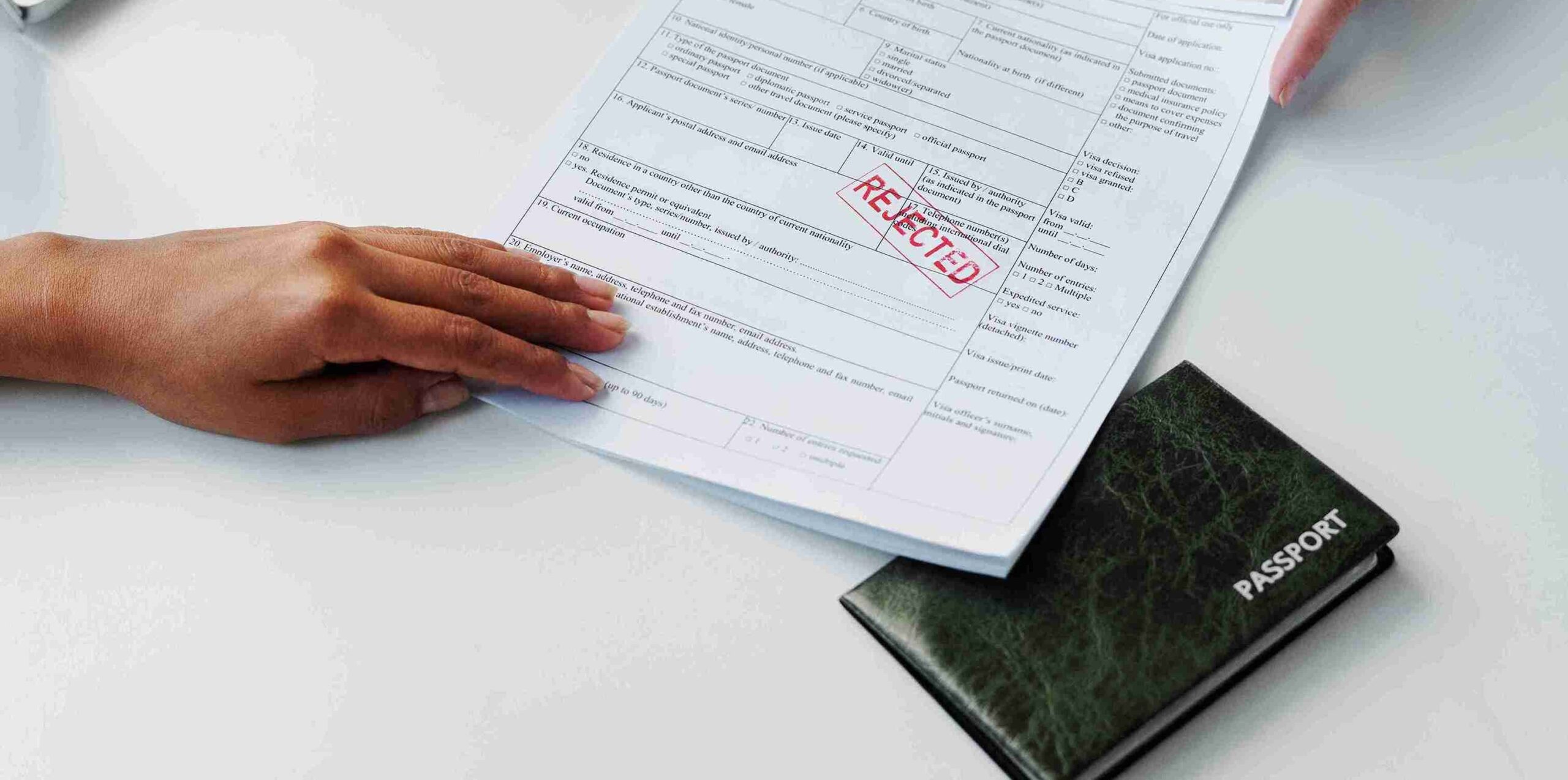 A person’s hands holding a printed application form stamped “REJECTED” in red, with a closed passport partially visible on the table beneath the papers.